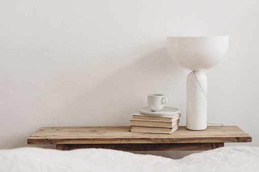 Cup of coffee on pile of books and modern marble geometric lamp. Vintage bench, table. White wall background. Scandinavian interior, bedroom. Selective focus, blurred bedding. Empty copy space.