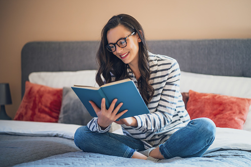 Happy young woman sitting on bed in her home and reading book.