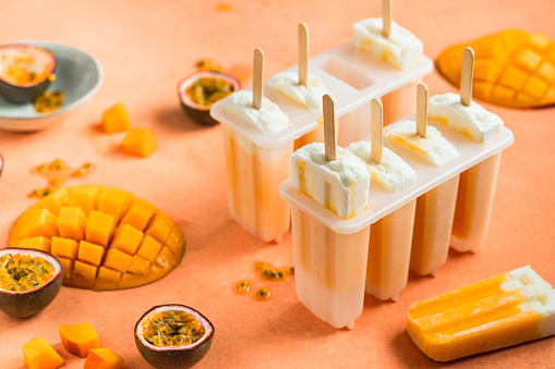 Homemade mix fruits ice cream lollies on orange table with lolly moulds and ingredients. Preparation of mango, passion fruit, popsicles in kitchen.