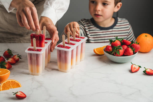 Woman and son making orange and strawberry ice pops Close-up of woman and son taking out ice pop from moulds in the kitchen. Mother and son preparing mis fruit ice cream on stick. Strawberries and oranges on table. homemade stock pictures, royalty-free photos & images