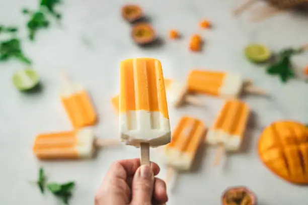 Close-up of a hand holding popsicle stick over a table. Making mango yogurt ice cream candy, with ingredients on the kitchen counter.