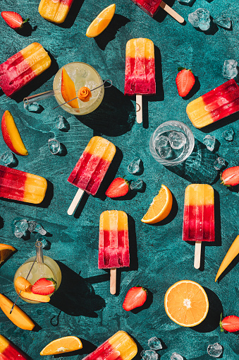 Strawberry, orange ice lollies with fresh fruit juice on blue table. Directly above shot of homemade ice lollies and juice over blue background.