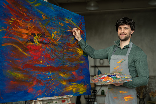 Portrait of male artist working on painting in studio. Male artist makes strokes with yellow paint.