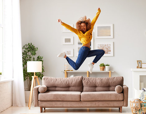 Enjoying freedom. Full length of overjoyed young afro american woman in casual wear jumping and dancing in living room, happy mixed race female teenager spending carefree time having fun alone at home