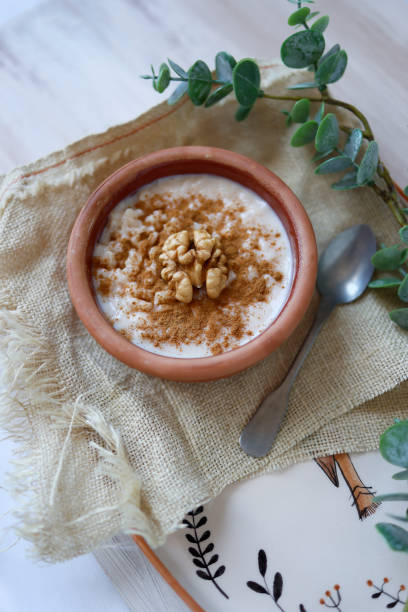 istock Rice pudding in a earthenware pot on ceramic tray on white tumbled wooden floor, Rustic lifestyle, Rice pudding decorated with cinnamon powder and walnuts, There are green tree branches around the tray 1318440512