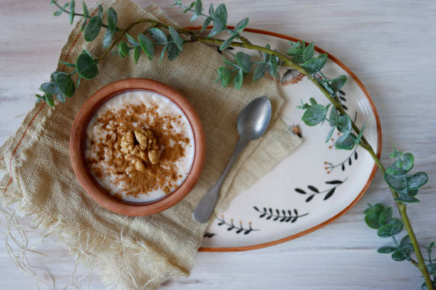 istock Rice pudding in a earthenware pot on ceramic tray on white tumbled wooden floor, Rustic lifestyle, Rice pudding decorated with cinnamon powder and walnuts, There are green tree branches around the tray 1318440655
