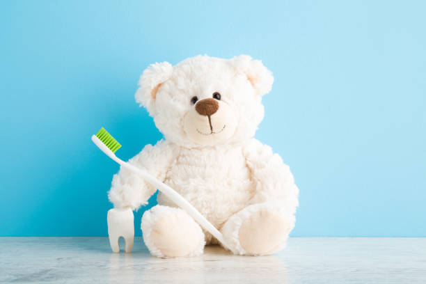 istock Smiling lovely teddy bear sitting with toothbrush and white tooth on table at light blue wall background. Pastel color. Closeup. Children teeth hygiene. Front view. 1318440740