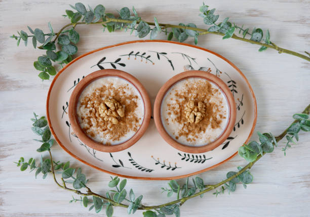 Rice pudding in two earthenware pot on ceramic tray on white tumbled wooden floor, Rustic lifestyle, Rice pudding decorated with cinnamon powder and walnuts, There are green tree branches around the tray