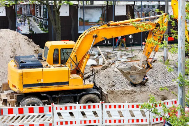 A heavy construction excavator works on a fenced-in construction site and digs a trench for urban utilities.
