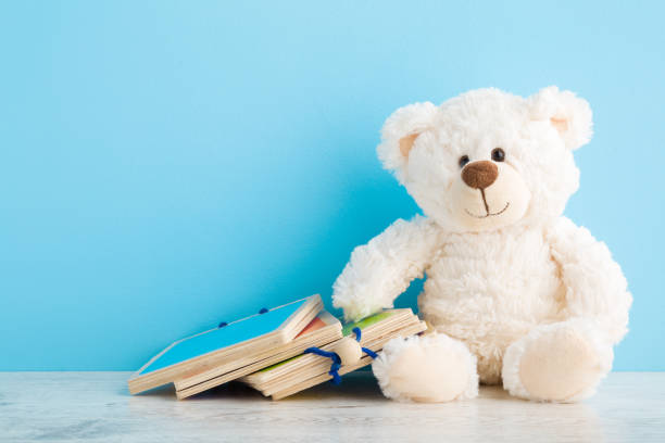 White smiling teddy bear with wooden books for baby development on table at light blue wall background. Pastel color. Closeup. Empty place for text. Front view.