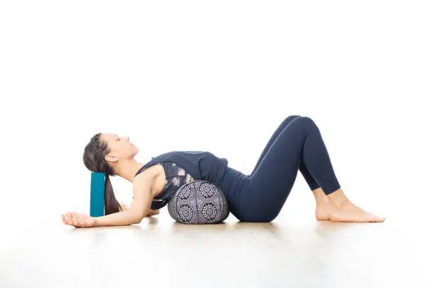 Restorative yoga with a bolster. Young sporty attractive woman in bright white yoga studio, lying on bolster cushion, stretching and relaxing during restorative yoga. Healthy active lifestyle.