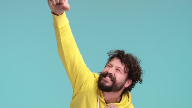 shocked cool hipster man with long beard and moustache making a surprised face, holding fists in the air, dancing and celebrating victory while yelling and having fun on blue background in studio