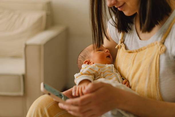 matka siedzi na łóżku i wpisując na telefon komórkowy, podczas gdy syn drzemki. - breastfeeding newborn mother baby zdjęcia i obrazy z banku zdjęć