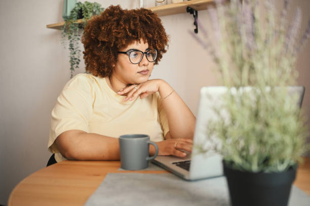 Attractive happy stylish plus size African black woman student afro hair in glasses studying online working on laptop computer at home office workspace. Diversity. Remote work, distance education. stock photo