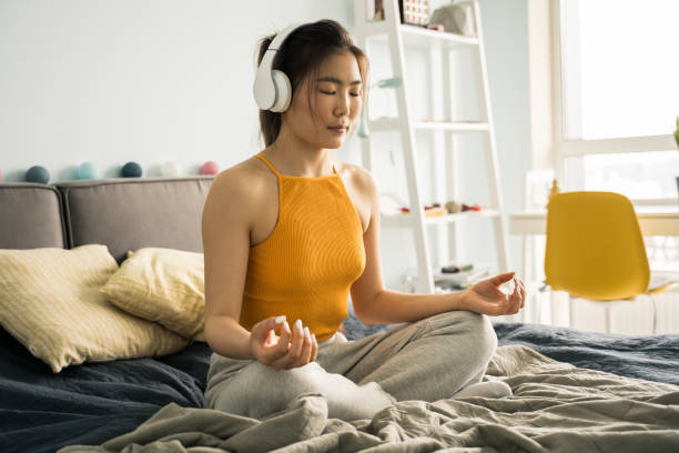 瞑想しながら、蓮の位置でベッドの自宅に座ってヘッドフォンを身に着けている女性 - lotus position audio ストックフォトと画像