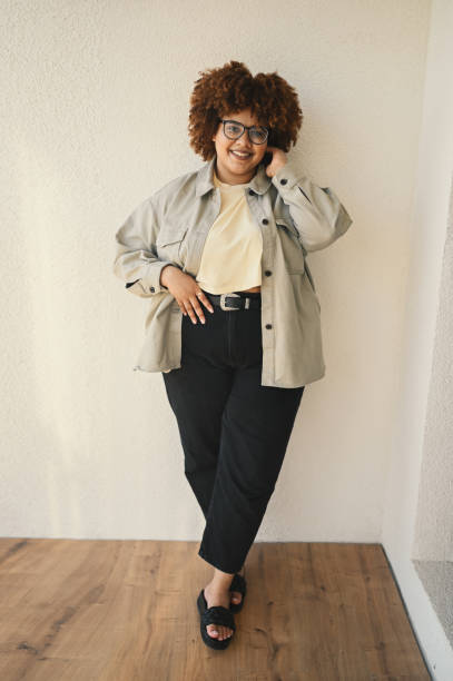 Beautiful happy smiling curvy plus size African black woman afro hair posing beige t-shirt, jeans, stylish glasses on beige background. Body imperfection, acceptance body positive diversity concept stock photo