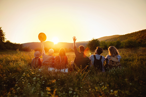 Group of young Caucasian friends, hikers, sitting on the field and waving at the sun.