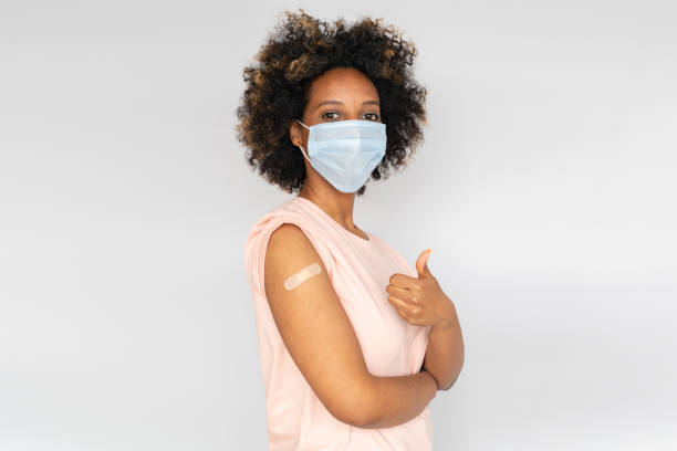 junge afrikanische amerikanische frau in medizinischer maske zeigt daumen nach oben und ihren arm mit bandhilfe nach coronavirus covid-19 injektion - thumbs up african descent teenager women stock-fotos und bilder