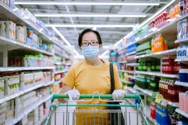 Asian Senior wearing face mask and rubber glove push shopping cart in suppermarket departmentstore. Girl choosing, looking grocery things to buy at shelf during coronavirus crisis or covid19 outbreak.