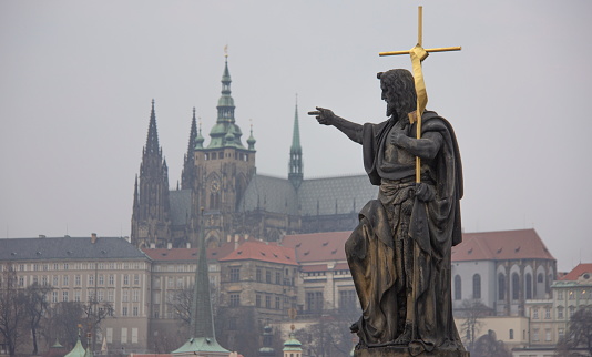 Sculpture of Saint John the Baptist bearing cross on Charles bridge with Prague Castle and  St. Vitus Cathedral in the background, Prague, Czech Republic