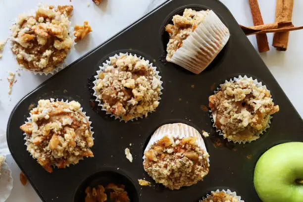 Stock photo showing elevated view of muffin baking tray containing homemade apple muffins, with streusel crumble topping, in paper cake cases on a marble effect surface. Home baking concept.