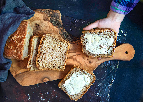 A child's hand holds a piece of bread with butter. Homemade whole grain bread with with seeds. Top view.