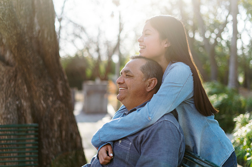 Beautiful latin teenage girl embracing father at park, looking away and smiling.