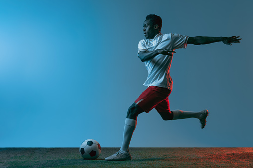 In action. Portrait of African man, male soccer football player in white red sports uniform isolated on blue background in neon. Concept of active life, team game, energy, sport. Copy space for ad.