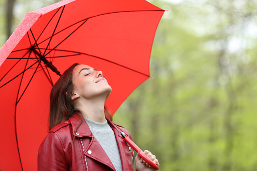 the model, who is 55 years old, has a red raincoat over a beret on her head.It's raining indoors.The season is winter.A large umbrella that plays pink in the model's hand.The model side angle is from the waist up.There are palm trees in the park