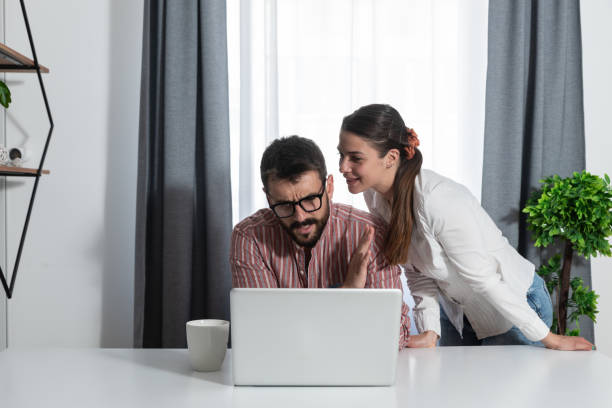 jovens colegas no trabalho onde uma mulher quer sussurrar para um colega fofoca que está acontecendo na empresa e ele mostra a ela com a mão que ele não está interessado porque ele trabalha em seu laptop - whispering couple discussion smiling - fotografias e filmes do acervo