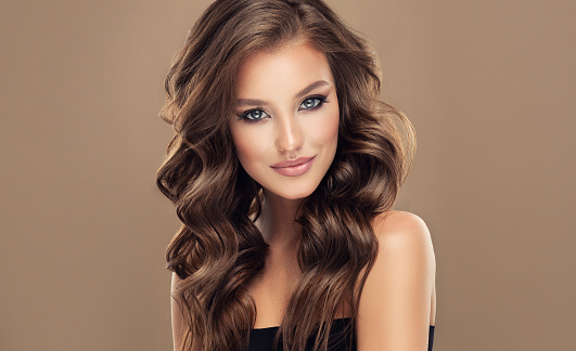 Portrait of young brown haired woman  with voluminous, shiny and wavy hair. Elegant makeup with black mascara and rose lipstick on her face. Beautiful model with long, dense and curly hairstyle.