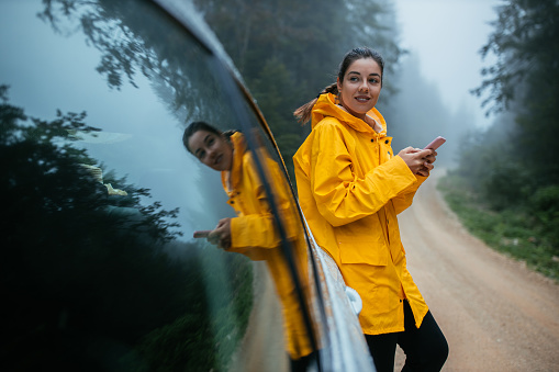 Young woman in yellow raincoat leaning at car on a mountain road while using phone