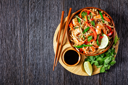 pad thai noodles stir-fry with prawns, carrots, red pepper, coriander and lime in a bowl on a bamboo tray with soy sauce and chopsticks, flat lay, free space