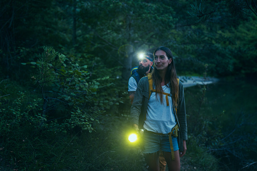 Photo of couple with backpacks holding flashlights and standing next to the lake.