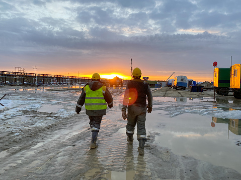 Uray, Russia-05.04.2021:Two oil workers in work clothes walk through the site along a muddy road to an oil rig in the background of the sunset. Heavy male work of drillers.