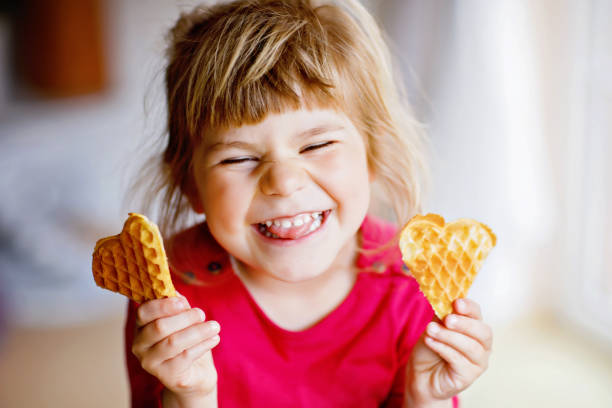 신선한 구운 심장 와플을 들고 행복한 작은 유치원 소녀의 초상화. 달콤한 비스킷 웨이퍼와 배고픈 유아 아이를 미소. 달콤한 설탕 벨기에 와플. - waffle breakfast food sweet food 뉴스 사진 이미지
