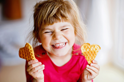 Portrait of happy little preschool girl holding fresh baked heart waffle. Smiling hungry toddler child with sweet biscuit wafer. Sweet sugar belgian waffles