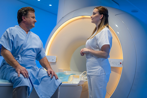 Mature man in hospital gown sitting on the MRI examination bed and talking with a female doctor in hospital.