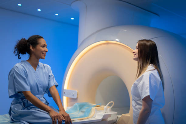 Mature woman talking with a doctor before MRI scan Mature woman in hospital gown sitting on the MRI examination bed and talking with a female doctor in hospital. radiologist photos stock pictures, royalty-free photos & images