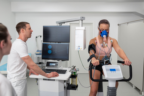 Male doctor observing the progress of a cardiopulmonary stress test taken by the female athlete riding a bicycle ergometer.