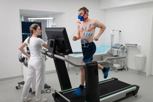 Male athlete running on a treadmill and taking a cardio stress Young male athlete running on a treadmill ergometer in his cardiopulmonary stress test overseen by a female doctor. stress test stock pictures, royalty-free photos & images