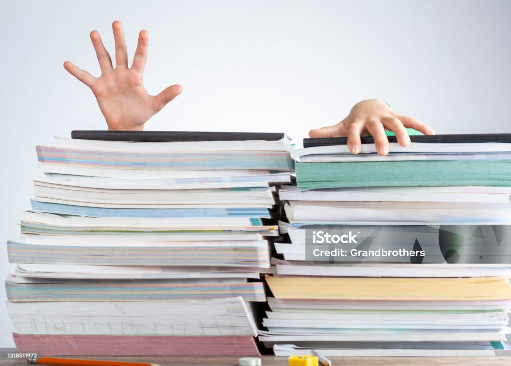 A child is behind a large pile of books. Demanding curriculum concept. Abstract concept image showing a young student behind a large pile of test prep books on a study desk. An overwhelming load. The kid is trying to escape by climbing onto the pile as if she is drowning Document Stock Photo