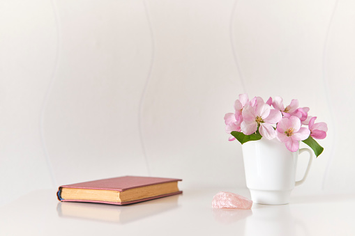 Interior composition. Gentle bouquet of pink apple tree flowers in a white coffee cup, pink mineral, small poetry book on a white coffee table against a light wall.