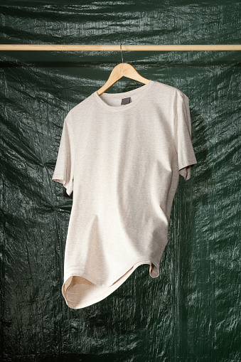 Beige t-shirt hanging on wooden coat hanger in front of green, plastic covered background