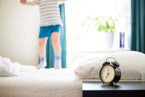 Just a woken up little child jumping on the bed with an alarm clock standing on the bedside table in front. Happy child jumping and playing on the bed.