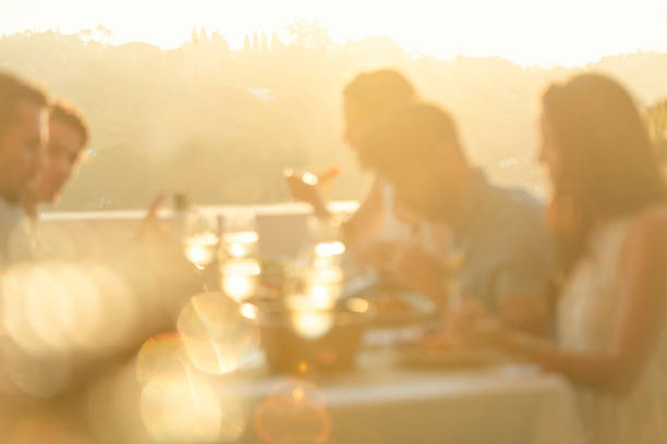 Friends enjoying a meal together on a table at sunset. Friends enjoying a meal together on a table at sunset. There are glasses of wine and other food on the table; everyone is having fun laughing and smiling. defocussed outdoor dining photos stock pictures, royalty-free photos & images