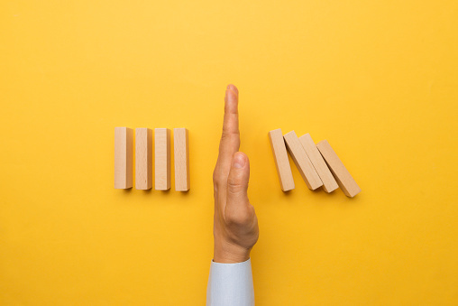 Hand is stopping chain reaction of dominos who are about to fall in front of a yellow background.