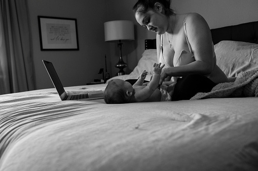 A mother fixes her baby’s cloth diaper on her bed, a computer is open next to her.
