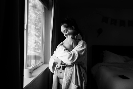A mother in a bathrobe cuddles her son next to a window in her bedroom