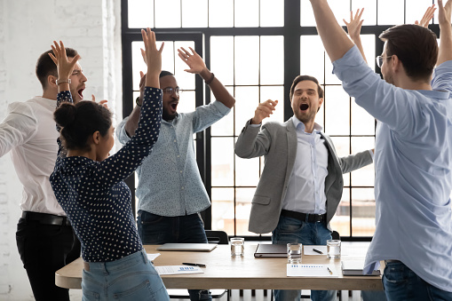 Happy excited millennial employees shouting for joy, raising hands for emotional high fives. Business team of overjoyed professionals cheering, celebrating good sales result, financial goal achieving